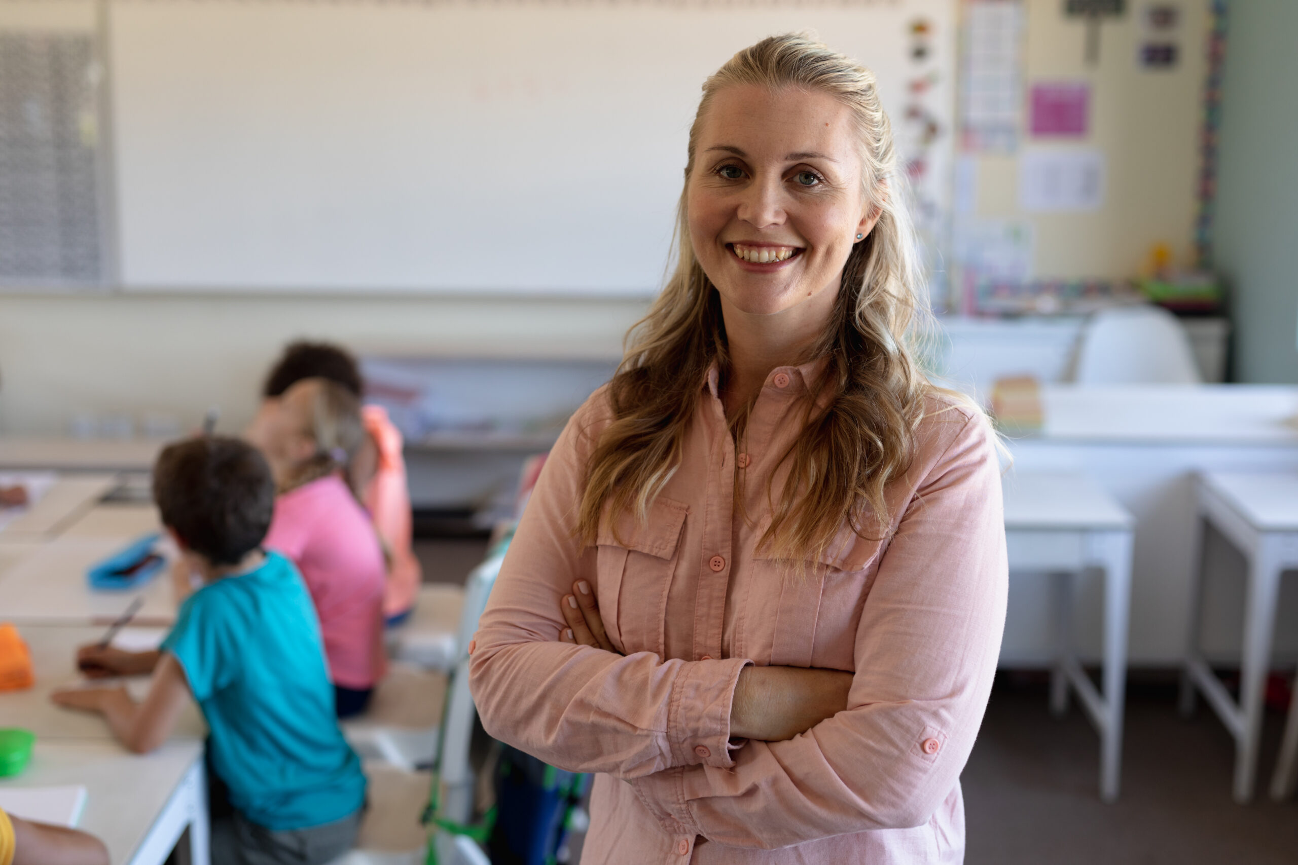 Portrait close up of a Caucasian female elementary school teacher with long blonde hair standing in a classroom with arms crossed, smiling to camera, with her pupils sitting at a table working in the background
